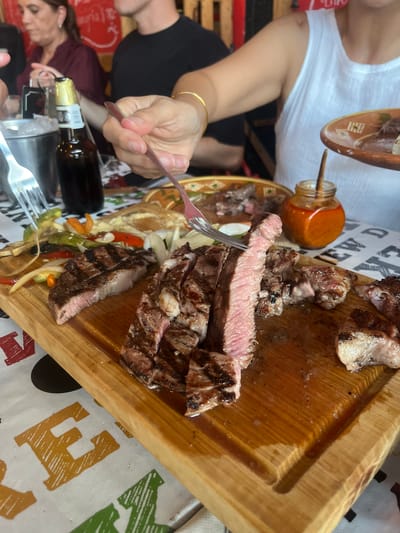 A medium-angle shot shows a person using a fork to pick up a slice of grilled steak from a wooden board laden with sliced steak and vegetables. A small glass jar filled with a reddish-orange sauce is nearby. Other items on the table include a plate, a bottle, and additional cutlery. The table is covered with a patterned tablecloth. The location may be San Francisco Totimehuacán, Mexico, though there is no explicit indication in the image.