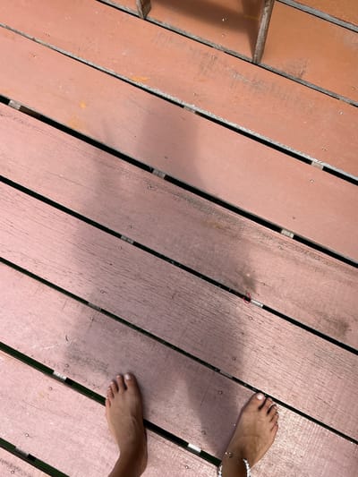 Two bare feet are visible on a wooden boardwalk. The feet are positioned at the bottom center of the image, with the left foot slightly closer to the camera. The wooden planks are reddish-brown and run diagonally from the top left to the bottom right. Gaps between the planks reveal a dark space underneath, possibly water. A faint shadow of a person or object is cast on the boardwalk from the top. A thin red thread or string is visible lying across one of the planks in the upper right quadrant of the image, near a small red circular object that appears to be a knot or a fastener. The scene is outdoors, likely in a warm climate, given the bare feet. The location is specified as Tambon Ko Panyi, Thailand, which is a floating village.