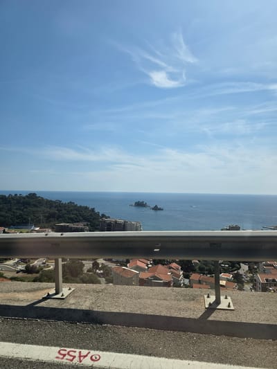 The image captures a panoramic view of the Adriatic Sea from a high vantage point, likely a road overlooking the coastal town of Brežine, Montenegro. The foreground features a weathered concrete barrier with a silver metal guardrail bolted securely onto it. The guardrail casts a distinct shadow on the concrete below. The road surface in the immediate foreground shows signs of wear and tear, with white and red markings visible, including the numbers "ASS".
Beyond the barrier, a densely forested hillside slopes down towards the sea. Nestled among the trees and on the lower slopes are numerous residential buildings with distinctive red-tiled roofs, characteristic of Mediterranean architecture. A few larger, more modern buildings are also present.
The sea stretches out to the horizon, appearing calm and a deep blue. A few small rocky islands are visible in the distance, with a small vessel positioned near them. The sky is a clear, bright blue, with wisps of white cirrus clouds streaking across it. The overall impression is one of a sunny day, with the coastline and sea bathed in natural light. No people are visible in the frame.