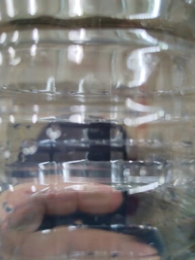 The image shows a blurry, close-up view of a hand reaching into a clear plastic bottle. The hand is mostly obscured by the bottle and distorted by the plastic and water. Reflected light creates bright streaks and circular patterns throughout the image. In the background, a dark, box-like object is visible through the bottle, possibly the base of a bottle cap or another object inside. The setting appears to be indoors, with indistinct background elements suggesting a domestic or possibly industrial environment. There is no discernible information about the location in Гожскі сельскі Савет, Belarus, within the image itself.