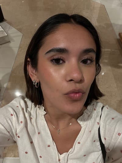 This is a vertical, slightly angled, close-up photograph of a young woman's face and upper chest, taken indoors. The subject has dark hair styled in a bob, dark eyebrows, and dark eyes. She is wearing subtle makeup, with emphasis on her eyelashes and lips, which appear glossy and slightly pink. A delicate gold necklace with a small cross pendant is visible around her neck, resting on her pale skin. She is also wearing a small, silver hoop earring in her visible ear.
Her clothing is a white or cream-colored blouse with a subtle pattern of small, reddish-pink dots. The blouse has ruffled detailing around the neckline and appears to be made of a textured fabric. A dark strap, possibly from a bag, is visible across her shoulder and chest.
The background consists of a tiled floor with large, beige and brown marble-patterned tiles, interspersed with lighter, square tiles. The lighting suggests an indoor environment, possibly a home or commercial space, with soft, diffused light that highlights the sheen on her skin. The overall mood is relaxed and casual. There is no visible text in the image. The location context provided is Orizaba, Mexico.
