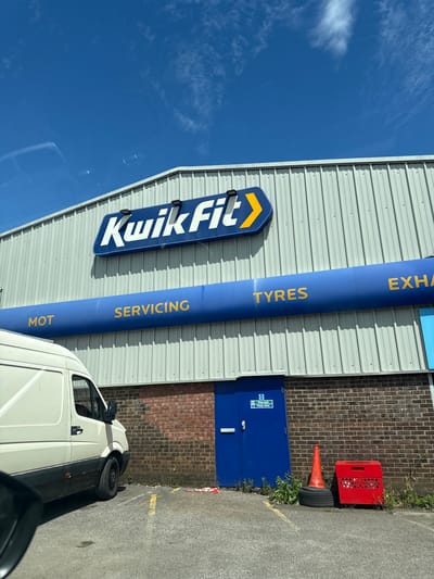 The image shows the exterior of a Kwik Fit building, a tire and auto repair shop, located in Nailsea, United Kingdom. The building has a corrugated metal facade with a prominent blue sign displaying the Kwik Fit logo. Below the logo, a large blue pipe runs horizontally, with text indicating services offered: "MOT", "SERVICING", "TYRES", and "EXHA". To the left of the building, a white van is parked. To the right of the van, a blue door with a "Fire exit - Keep clear" sign is visible, set into a brick wall. In front of the door, a traffic cone is placed on a tire, and a red plastic crate is nearby. The sky above is blue with scattered white clouds. The ground is asphalt.