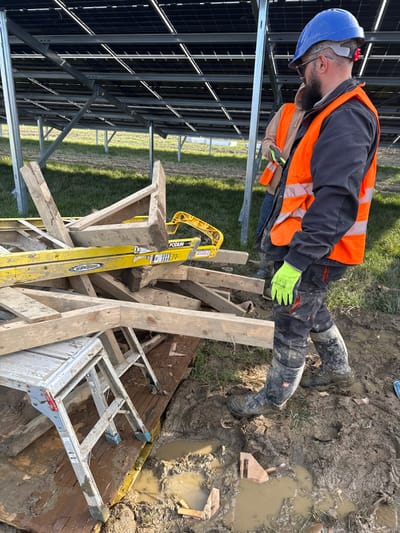 The image depicts an outdoor construction or maintenance site at Baldurgan Hill, Ireland. In the foreground and mid-ground, a heavily muddy ground surface features numerous puddles of brown water and distinct footprints. A pile of wooden planks and beams lies haphazardly on the ground, partially obscuring a yellow "Werner Podium" ladder and a silver aluminum step stool/work platform, all coated in mud.
Two male individuals are present. The primary subject, positioned in the mid-ground on the right, faces towards the left and downwards. He wears a blue hard hat with safety glasses, a dark jacket, an orange high-visibility vest with reflective stripes, black work pants, and black muddy work boots (brand "ELTEN"). Bright yellow/lime green work gloves are visible on his hands. A beard is visible on his face. Behind him, partially obscured, a second male individual is visible, wearing a light brown jacket and an orange high-visibility vest.
In the background, a large solar panel array structure extends horizontally across the frame. Rows of dark solar panels are mounted on silver-colored metal frames, supported by vertical posts embedded in a grassy field. The scene is illuminated by natural daylight.
