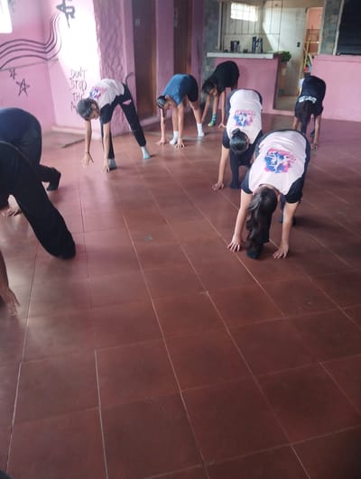 The image depicts a group of young girls engaged in a dance or gymnastics session in what appears to be a studio. They are wearing athletic attire, primarily black leggings and t-shirts, with some wearing shirts with colorful graphic designs. All the girls are in a bent-over position, with their hands reaching towards the floor, indicating a stretching or warm-up exercise. The environment features a pink wall with graffiti that includes musical notes and text. The floor is tiled in a reddish-brown color. The lighting suggests an indoor setting, possibly a community center or a dedicated dance studio. The overall scene captures a moment of physical activity and practice among the participants. The location is identified as El Espinal, Venezuela.