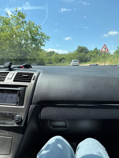 The image is taken from inside a car, focusing on the dashboard and the view through the windshield. The dashboard is black and gray, with visible air vents and a car stereo displaying "87.50 MHz" and buttons for "TRACK," "VOL," "A/C," "TEMP," and "DUAL." The driver's leg is visible on the right, wearing light blue jeans. Outside the car, a road winds through a green, wooded area under a bright blue sky with scattered clouds. A road sign indicating a curve to the right is visible. Another car is ahead on the road. The image provides no explicit information to confirm that the location is in the city of Levski, Bulgaria.
