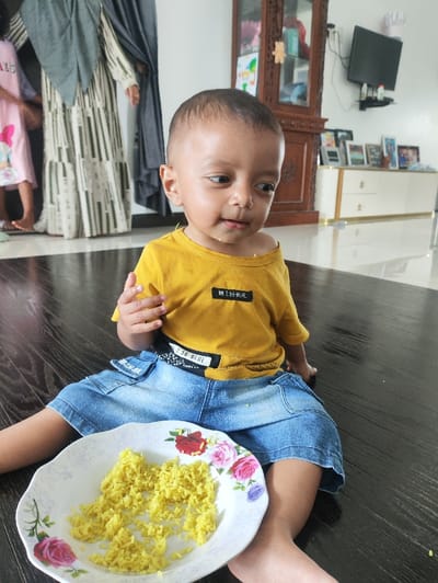 An indoor scene in Meunasahbaro, Indonesia, depicts a young child seated on a dark, reflective floor with a plate of yellow rice. The child, with short dark hair and light brown skin, wears a yellow t-shirt with "THE BEST BLUE" text and denim shorts, appearing to have food residue around the mouth and looking towards the right. A white plate with a floral rose pattern (pink, red, purple) rests on the floor in front of the child. In the blurred background to the left, the lower portions of two individuals are visible; one wears a pink garment, and the other a long-sleeved white and green striped dress under a dark grey head covering. To the right, a dark wooden display cabinet with glass doors stands near a low, light-colored cabinet topped with multiple framed pictures. A black flat-screen television is wall-mounted above the lower cabinet, and a white chef's hat decoration is affixed to the wall beside it. The room features white walls and a polished dark floor.