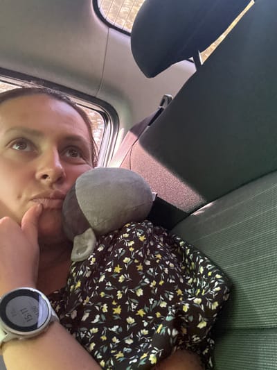 An adult female is depicted in the passenger seat of a car, located in Simakovo, Russia. She has dark hair and a pensive expression, looking upwards and to the right of the frame, with her right index finger touching her chin. She is wearing a dark garment with a yellow and white floral pattern. On her left wrist, a white smartwatch displays "15:11" and "11360." A grey, plush stuffed animal rests against her, partially covered by her dress. The vehicle's interior features dark grey/green fabric seats and a light grey headliner. Bright sunlight enters from a window on the right, illuminating parts of the interior and casting shadows. The view through the window is blurred, showing indistinct outdoor scenery.