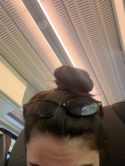 The image shows a close-up of a person's head from a low angle, looking upwards. The person is wearing dark-rimmed sunglasses positioned on their forehead, atop a dark hair bun. The background consists of a ceiling with linear patterns and lighting, suggestive of interior lighting in a vehicle or building. The ceiling panels are off-white with black linear indentations, and strip lighting runs along the length of the ceiling. A round, white smoke detector is visible on the ceiling to the left of the frame. The lighting is warm and appears to be from fluorescent or LED fixtures. A portion of a dark-colored seat is visible on the right side of the image. The overall impression is of an indoor environment, possibly public transportation given the ceiling design. The location is identified as Vejprnice, Czechia, though no specific landmarks or urban features are discernible within the image itself to confirm this.