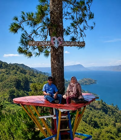 A man and a woman are seated on a colorful wooden platform elevated by a tree, overlooking a vast blue lake and verdant hills under a clear sky. The man, on the left, wears a blue button-down shirt, jeans, and a black baseball cap. He holds a mobile phone in his lap and has sunglasses on his head. The woman, on the right, is dressed in a patterned headscarf and a long-sleeved top with matching trousers. They are both looking towards the camera. A wooden sign attached to the tree reads "BUKIT INDAH SINIR MARIDALINUS" with a heart graphic. The platform is supported by a structure of brightly painted wooden beams. The background features a dense, green forest, the expansive lake, and distant mountains. This location is identified as Bukit Indah Simarjarunjung, a popular tourist spot in Parik Sabungan, Indonesia.