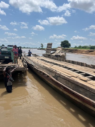 A group of people are gathered around a damaged wooden ferry on a muddy river. A green SUV is parked on the ferry's deck, suggesting an attempt to transport it across the river. The ferry appears to be made of multiple connected wooden boats, some of which are significantly broken and dilapidated. The riverbank in the background is lined with vegetation, and trees are visible further inland. The sky is blue with scattered white clouds. The scene is set in Nemnei, Nigeria.
