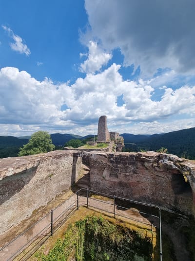 The image shows the ruins of a castle on a hilltop overlooking a forested valley under a bright blue sky with scattered white clouds. The foreground features the stone walls of the castle, weathered and overgrown with moss and grass. A metal railing guides visitors along a path next to a steep drop. In the background, the remnants of a tower and other stone structures stand against the backdrop of rolling green hills. The scene is set in the morning or afternoon, as indicated by the strong sunlight casting shadows. This is the Dahner Schlossberg, a historical landmark in Dahn, Germany, known for its ancient fortifications and scenic views. No people are visible in the image.