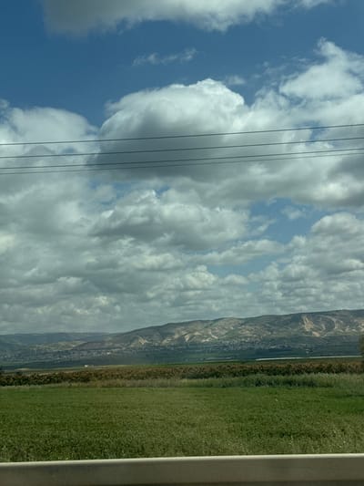 The image presents a wide-angle landscape view captured from a vehicle, looking across an agricultural plain towards distant hills under a partly cloudy sky within the Emek HaMaayanot Regional Council, Israel. The upper half of the frame shows a blue sky dominated by numerous white and grey cumulus clouds, with clear blue patches visible between them. Four thin, dark power lines stretch horizontally across the upper-middle section of the image. The immediate foreground is occupied by a light-colored, horizontal barrier, likely a vehicle's dashboard or a road guardrail. Beyond this, a vast expanse of vibrant green cultivated fields fills the lower-mid ground. Farther back, a band of denser, darker vegetation, possibly mature crops or natural flora, precedes a valley floor where small clusters of buildings, indicative of settlements, are discernible. The background features a continuous range of light-colored hills or mountains, displaying varying patterns of vegetation and exposed earth. No individuals or specific activities are observed.