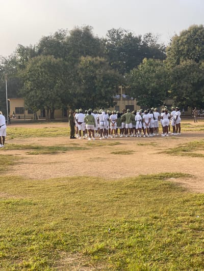 The image depicts a group of individuals, appearing to be cadets or trainees, in an outdoor setting. They are predominantly dressed in white shorts and white t-shirts, with some individuals also wearing green shirts and caps. A single person in military camouflage attire stands slightly apart from the larger group. The individuals are arranged in rows, facing away from the camera, in what looks like a parade ground or open field. The ground is a mixture of dirt and sparse grass. In the background, there are trees and a building with a light-colored facade. The overall scene suggests a formal gathering or training exercise. The location is identified as Ise- Ekiti, Nigeria.