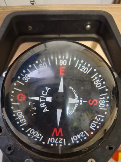 The image shows a close-up of a black-cased marine compass. The compass rose has markings for degrees and cardinal directions (N, S, E, W). "S" (South) is marked in red at 180 degrees. The compass face is labeled "Riviera Genova, Italy." The brand "ARTICA" is also visible. The compass appears to be mounted within a black, bracket-like structure. This image does not provide any information about the city of Warffum, Netherlands.