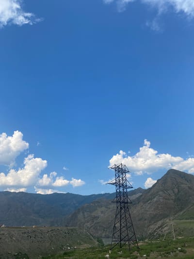 The image captures a vast, clear blue sky with scattered white cumulus clouds. In the foreground, a tall, dark metal power line tower stands prominently. Beyond the tower, a range of imposing, rugged mountains dominates the middle ground, their slopes textured with shadows and lighter hues. A winding river is visible in a valley between the mountains on the left. The bottom of the frame shows a grassy, rocky hillside. A few small buildings, possibly houses, are nestled on a shelf of land in the lower left portion of the mountain range. The scene appears to be set in a rural, mountainous region, possibly near Inya, Russia, given the characteristic landscape.