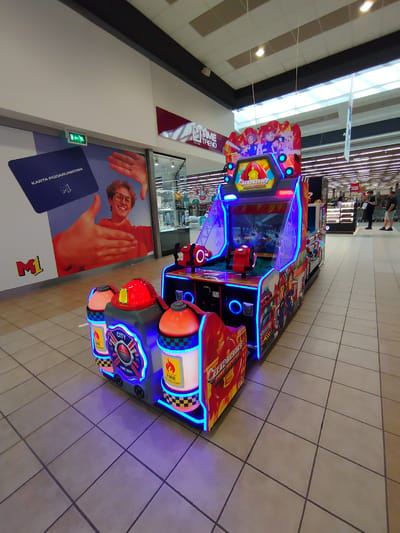 In the city of Marki, Poland, at a M1 shopping mall, an arcade game machine with a fire rescue theme is displayed. The machine features bright neon lights and graphics of cartoon firefighters and fire trucks. The game is titled "Real Heroes: Fire Rescue." In the background, store displays and ceiling lights of the mall are visible, with a few shoppers in the distance. The floor is tiled with a light-colored ceramic material.