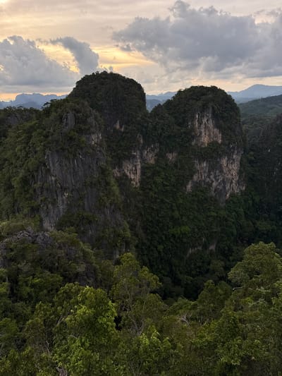 The image depicts a dramatic landscape of karst mountains in Ban Khao Tang, Thailand, likely during a cloudy or twilight period. The foreground is dominated by lush green trees, with their leaves forming a dense canopy that partially obscures the view of the mountains beyond.
The main subjects are two imposing, steep-sided mountains that rise from the surrounding jungle. These mountains are characterized by sheer rock faces, displaying textures of gray and brown stone, with vertical striations and crevices. They are heavily vegetated, with dense green foliage clinging to their slopes and summits, softening their rugged appearance.
In the distance, additional layers of mountains recede into a hazy, muted landscape, suggesting a vast, undeveloped area. The sky above is filled with a mixture of clouds, ranging from dark, heavy formations to lighter, illuminated patches where the sun may be breaking through or setting. The overall impression is one of natural grandeur and a remote, untamed environment. No people or specific man-made objects are visible in the scene.