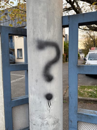 Captured through a blue metal fence, a grey concrete post bearing black spray-painted graffiti in the shape of a question mark and two short, parallel lines is prominently visible. Behind the fence, an out-of-focus street scene includes a light-colored building with windows, a white van, bare trees, and greenery. The photo was taken in Orsay, France.