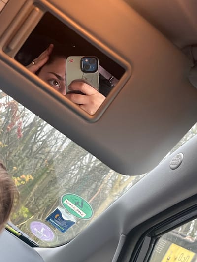 The image depicts the interior of a vehicle located in Lonate Pozzolo, Italy. The primary subject is a sun visor mirror, angled downwards from the car's ceiling, reflecting a person. This individual's face, specifically the eyes and forehead, and both hands are visible in the reflection. One hand, adorned with a gold ring, holds a light green-cased smartphone featuring a dual-camera system, apparently capturing the reflection. The other hand rests on the individual's head.

The vehicle's gray textured ceiling and A-pillar are visible, with an "SRS CURTAIN AIRBAG" label present on the A-pillar. The windshield displays three affixed stickers: a prominent green oval sticker with white Arabic text translating to "Private Parking," a purple oval sticker partially showing "MILANO" text, and a blue/white rectangular sticker containing a QR code. Through the slightly soiled windshield, a densely treed outdoor environment is visible, characterized by bare branches and sparse foliage, with some reddish hues, indicating an autumn or winter setting under diffused lighting. A portion of brown hair is visible in the lower-left foreground, slightly out of focus.