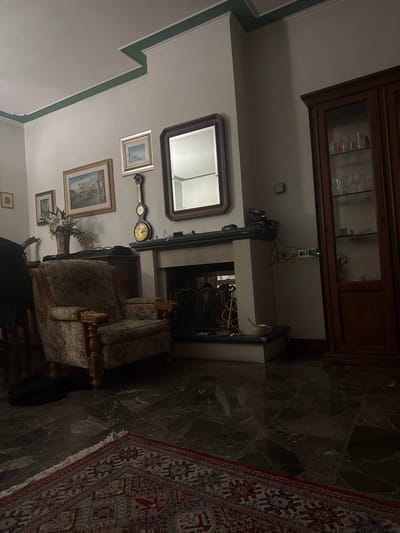 The image depicts a dimly lit interior space, possibly a living room, with a mix of traditional and antique-style furnishings. A dark wooden cabinet stands tall, with glass shelves displaying various objects. Below the cabinet is a fireplace with a stone mantelpiece. On the wall above the fireplace is a mirror. The wall also features framed paintings and a barometer/clock. An armchair sits near the fireplace. A rug with a geometric pattern is spread on the floor.
Without additional information, determining the specific location in Perugia, Italy is not possible.