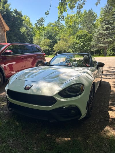 An outdoor, eye-level, medium shot captures a white convertible sports car parked on a gravel lot. The car, facing slightly to the left, is illuminated by sunlight, casting shadows from the surrounding trees. To the left of the sports car, a red SUV is partially visible. The background consists of lush green trees and a clear blue sky, suggesting a sunny day. The scene is set in Bouctouche, Canada.