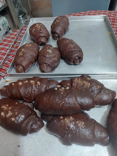 A close-up, eye-level shot shows two metal baking trays filled with chocolate-colored bread rolls. The rolls on the top tray are elongated and appear to be twisted, with some sprinkled with coarse sugar. The rolls on the bottom tray are crescent-shaped and also sprinkled with sugar. The trays are placed on a surface covered with a red and white checkered cloth, and a wire rack is visible to the left. In the background, shelves and industrial equipment suggest a bakery or commercial kitchen setting. The overall impression is of freshly baked goods prepared for sale. No specific location in Jelutung, Indonesia, can be determined from the image.