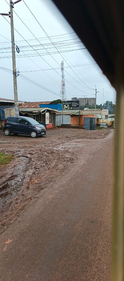 A dark compact car sits beside a muddy street with deep tire tracks. Several small buildings and shops line the street, with rusty roofs. A sign advertising "Easters Lounge" is visible. In the background, there's a tall cell tower and a high-rise building under construction. The day appears overcast, with visible power lines. The photograph seems to be taken from within a vehicle, with a portion of the frame obstructed.