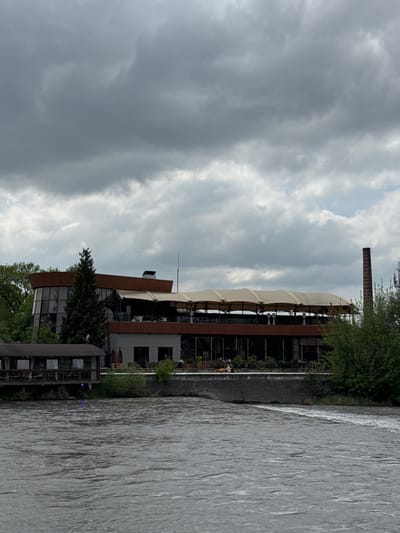 The image depicts a multi-story building situated beside a river. The building has a mix of modern architectural styles, including glass walls, metal framework, and a tent-like roof structure on one section. A tall, brick chimney-like structure stands nearby. The river appears wide and flowing, and its surface shows rippled textures. The sky is overcast, with dark, heavy clouds dominating the scene. Vegetation is visible around the building and along the riverbank, including trees and shrubs. The details provided do not confirm the location in the city of Zamankul, Russia. More forensic information is required.
