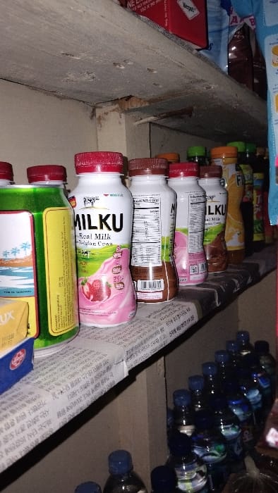 The image shows a shelf in a small shop, possibly a convenience store, located in the city of Panjalu, Indonesia (latitude: -7.142, longitude: 108.278). The shelf is made of wood and is covered with a layer of dust and debris. There are various products arranged on the shelf, including a variety of milk products, bottled water, and other miscellaneous items. The milk products are in plastic bottles with red lids, while the bottled water is in clear glass bottles with blue lids. There are also various products, some in boxes and some in plastic containers, on the shelf. The image shows a close-up view of the shelf, and the products are not clearly identifiable. There are no people in the image. The environment appears to be dimly lit. The image appears to have been captured in a vertical orientation.