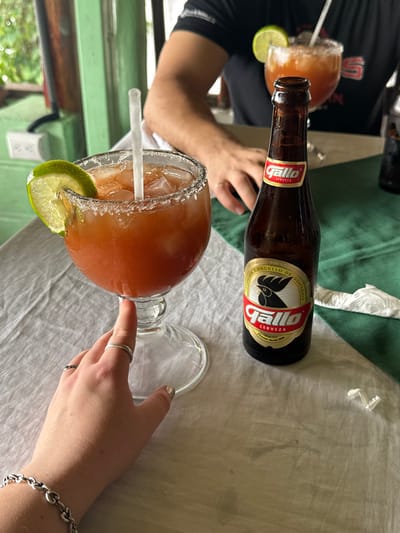 A table setting featuring two Micheladas with salted rims and lime wedges, alongside a bottle of Gallo beer. A hand with rings rests on the stem of one glass, with a silver bracelet visible on the wrist. Another person's arm is partially visible. The table has a green accent and a light-colored tablecloth with some green fabric.