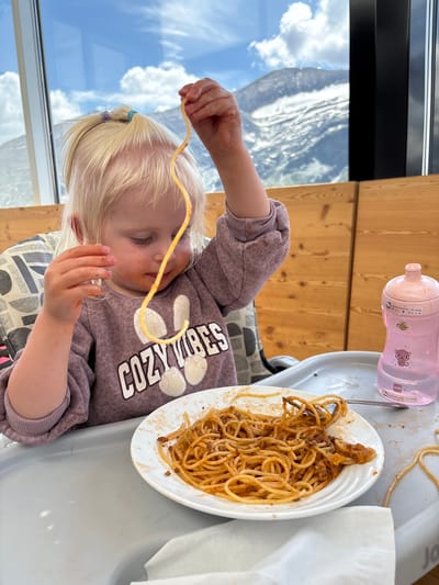 A young child with blonde hair pulled into pigtails with blue and purple ties is sitting in a high chair and eating spaghetti. The child is wearing a purple sweatshirt with "Cozy Vibes" printed on it in white letters. A plate of spaghetti is in front of the child, and they are holding a strand of spaghetti up to their mouth. To the right of the plate is a pink sippy cup. In the background, there is a window with a view of mountains and a cloudy sky. The scene appears to be in a restaurant in Heiligenblut am Großglockner, Austria, as indicated by the background scenery which is characteristic of the region.