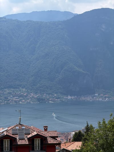 The image captures a scenic view of Abbadia Lariana, Italy, overlooking Lake Como. In the foreground, the terracotta-tiled roof of a red house is visible, equipped with solar panels and a television antenna. Beyond the house, the expansive blue waters of Lake Como stretch out, with a white boat leaving a wake. In the middle ground, a picturesque town with red-tiled roofs and white buildings lines the shore. The background is dominated by lush green mountains, their slopes covered in dense vegetation. The sky is partly cloudy.