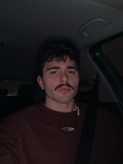A person with dark, curly hair and a drawn-on mustache looks towards the camera. They are lying in what appears to be the back of a car, with the vehicle's interior visible. The person is wearing a dark brown sweater. The lighting is dim, and the image is slightly rotated counterclockwise.