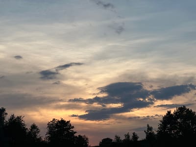 This is an image of a cloudy sky at sunset. The clouds are a mix of dark gray and lighter white, with streaks of orange and yellow from the setting sun. The top of the image is silhouetted by trees. The overall mood of the image is peaceful and serene, with a hint of drama from the colorful sky. The image was taken in Halfing, Germany.