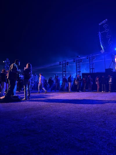 The image shows a nighttime scene at what appears to be an outdoor concert or festival in Coachella, California, United States. There is a crowd of people standing in front of a stage that is equipped with lighting rigs. The stage lights are casting a blue glow. The ground appears to be grass. Some people in the foreground are looking at their phones.