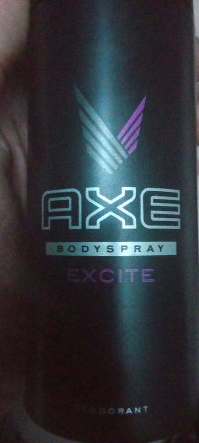 The image shows a close-up of a dark-colored Axe bodyspray can, specifically the "Excite" scent. The can has a matte finish with metallic-style lettering and a graphic logo that appears to be a stylized wing. The text "AXE" is prominently displayed in large, outlined letters, followed by "BODYSPRAY" in a smaller font, and then "EXCITE" in a larger, purple, outlined font. The bottom of the can also shows the word "DEODORANT" in faint lettering. The background is dark and appears to be indoors, with some lighting creating reflections on the can. There is no indication of any specific location in Vozdvijenka, Russia, or any people or actions.