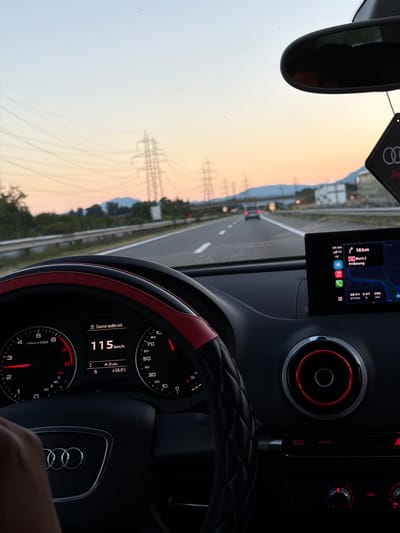 The image shows the interior of a car, specifically the dashboard and steering wheel, as seen from the driver's perspective. The car is on a highway, with a sunset visible through the windshield. The dashboard displays the car's speed at 115 km/h and the outside temperature at +16.0°C. A navigation screen shows a route to Bern/Fribourg, indicating a distance of 16 km. The car appears to be an Audi, based on the emblem on the steering wheel and the design of the air vents. Electrical pylons and trees line the side of the highway. The scene is set during dusk, with a warm, orange hue in the sky. The location is identified as Yvorne, Switzerland.