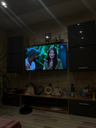 The image shows the interior of a home, likely in Popintsi, Bulgaria. The focal point is a television displaying a scene with a woman and a man. Above the TV are dark-colored cabinets. To the right is a shelving unit displaying various decorative items, and a floor lamp is positioned nearby. A vacuum cleaner is visible on the floor, and a plant is located in the lower portion of the photo. The scene is dimly lit. The flooring appears to be laminate.