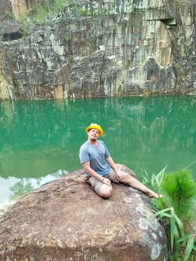 A man with a beard and a yellow hard hat is sitting on a large, rough-textured boulder. He is wearing a blue t-shirt and khaki shorts. His left leg is bent, with his knee up, and his right leg is extended, with his foot resting near some green vegetation. Behind him, a vibrant green body of water is visible, reflecting the rocky cliff face that rises steeply behind it. The cliff is layered and textured, with streaks of different colors and patches of green growth. The overall scene appears to be a quarry or a similar natural setting. The location is identified as Lán Tranh city in Vietnam.