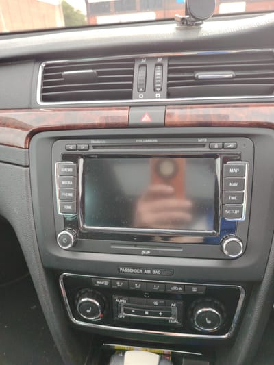 The image displays the central dashboard console of a Škoda vehicle, indicated by the "Škoda Auto" branding on the infotainment system. The scene is depicted as being located in Peterborough, United Kingdom. The upper section of the dashboard features a set of horizontally louvered air vents with chrome surrounds. To the left of these, two vertical scroll-wheel controls are present. A small, black, multi-jointed mount is attached to the top surface of the dashboard, near the windshield. A horizontal panel with a dark wood-grain effect runs across the mid-section of the dash, separating the air vents from the infotainment unit. Below the central air vents, a red triangular hazard warning button is visible.
The primary subject is the "COLUMBUS" branded infotainment system, featuring a large, rectangular touchscreen display. The screen is reflective, showing a blurred image of a human hand holding a rectangular object, likely a smartphone, with an orange or brown case. To the left of the screen, there are four vertically stacked buttons: "RADIO", "MEDIA", "PHONE", and "TONE". To the right, another stack of four buttons reads: "MAP", "NAV", "TMC", and "SETUP". Above the screen, several smaller buttons include left and right arrows, "MP3", and symbols for information and mute/volume. A rotary knob with a power/on-off symbol is located on the bottom left of the infotainment unit, and a matching rotary knob is on the bottom right. An SD card slot is visible directly below the screen. Below the infotainment system, a "PASSENGER AIR BAG OFF" indicator strip is present.
The lowest section of the console houses the climate control panel. This panel includes various buttons for functions such as "MAX" defrost, front and rear defoggers, air recirculation, "AC", "AUTO", "DUAL" zone control, and fan speed adjustments (plus and minus). Two large rotary temperature control knobs are positioned at the bottom, each featuring a seat heater symbol (three wavy lines) and a red indicator dot, suggesting active heating functionality. The dashboard surfaces, particularly the screen, exhibit minor dust and smudges. The overall interior color scheme is dark, with silver-colored chrome accents and a wood-grain trim providing contrast. No individuals are directly visible, only the reflection of a hand on the screen.