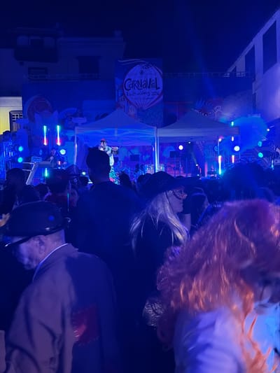 This image depicts a nighttime carnival event occurring in Icod de los Vinos, Spain, specifically referencing "Carnaval Icod de los Vinos 2026" on a large banner spanning a building facade in the background. The scene is dominated by vibrant blue and purple artificial lighting emanating from stage equipment.

In the midground, a raised stage is visible, featuring at least two white canopy tents. A male performer wearing a light-colored outfit and a cap is actively singing into a microphone, positioned centrally on the stage. Additional figures and musical equipment are dimly discernible around him. The stage is flanked by vertical LED light bars displaying multiple colors and various spotlights. Large, stylized decorative elements, including what appears to be a large shell-like structure, are visible on the left side of the stage.

The foreground is densely populated with a crowd of individuals, largely facing towards the stage. On the left foreground, a male individual wearing a dark bowler hat, glasses, and an earring is seen from the back-left shoulder, clad in a light-colored jacket with a distinct red square patch stitched onto the back. On the right foreground, a person with long blonde hair wears a dark hat and a prominent, ornate, sparkling eye mask. To the extreme right, out of focus, a person with voluminous reddish-orange hair is partially visible. The crowd appears to be engaged in observing the performance. The overall environment is an urban setting at night, with dark building structures providing the backdrop to the illuminated event.