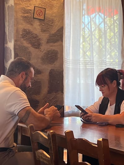 The image captures an indoor scene within an establishment in Civitavecchia, Italy, likely a restaurant or café. Two individuals are seated opposite each other at a glossy, reddish-brown wooden table.

On the left, a male subject with short dark hair, a beard, and glasses, wearing a white polo shirt, is focused on a black smartphone held horizontally with both hands. A tattoo is visible on his right upper arm. On the right, a female subject with reddish-brown hair, styled with bangs and a ponytail, and wearing glasses, a white t-shirt, and a dark vest, is focused on a black smartphone held vertically with both hands. Her phone has a visible ring stand accessory on its back. No direct interaction between the two individuals is evident, as both are independently engaged with their devices.

The environment includes a textured wall behind the male subject, appearing to be rough stone or a stone-like finish with irregular dark brown and lighter beige/grey patterns. A square "no smoking" sign, featuring a red circle with a diagonal line through a cigarette icon, is mounted on this wall. To the right, a window covered by sheer white curtains reveals green foliage and a red structure, possibly a tiled roof or awning, outside. Partially visible wooden chairs with a rustic design are positioned in the foreground at the table.
