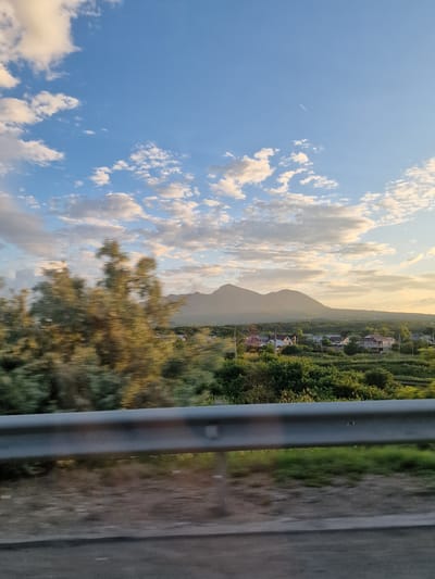 The image depicts a landscape scene with a road in the foreground, separated by a metal guardrail. Beyond the guardrail, a village is nestled among green trees and rolling hills, leading to two distinct mountains in the background. The sky above is a clear blue, dotted with scattered white clouds, and a faint contrail from an aircraft is visible. The lighting suggests it is late afternoon or early evening, with a warm glow illuminating the scene. The village appears to be located in the vicinity of Mount Zmeyka, in Zmeika, Russia, given the characteristic double-peaked mountain formation.