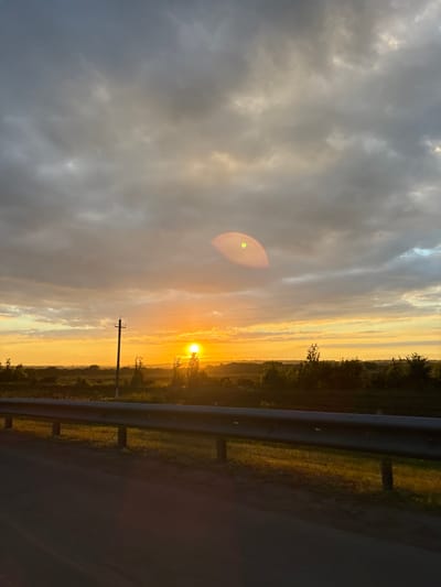 The image depicts a sunset over a rural landscape, likely in Im. Tattsika, Russia, given the provided context. The sun is low on the horizon, casting a warm orange glow that illuminates the sky and a field. Dark, scattered clouds dominate the upper portion of the sky, with some lighter patches where the sun's rays break through. A solitary utility pole stands prominently in the middle ground, with power lines stretching across the frame. The foreground features a guardrail along what appears to be a road, with the road surface itself in shadow. Trees and vegetation line the horizon, silhouetted against the bright sky. A faint lens flare is visible in the upper right quadrant of the image. There are no people visible in the scene.