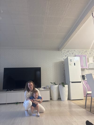 A woman holds a toddler in her arms, both seated on a light-colored floor in what appears to be a home interior, possibly in Nestiukovo, Russia. The room includes kitchen appliances such as a refrigerator, as well as a TV and shelving unit. Wallpaper with a patterned design is visible on the walls.