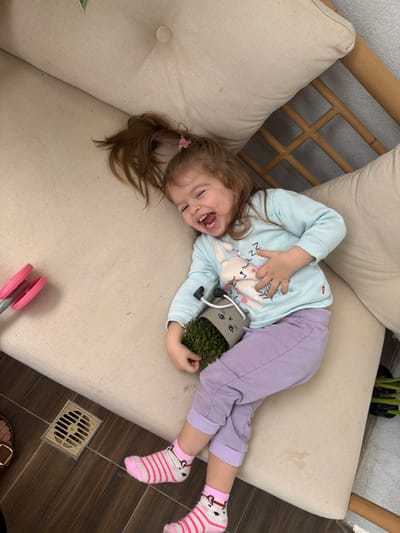 The image captures a young female child, estimated to be between two and four years old, positioned supine on a light beige cushioned bench in an outdoor or semi-outdoor setting within the city of Harish, Israel. The child has light brown hair gathered into a high ponytail secured with a pink accessory. She is dressed in a light blue long-sleeved shirt with a white rabbit graphic, light purple pants, and pink and white striped socks featuring an animal motif. Her mouth is wide open, and her eyes are squinted, indicative of a strong vocalization or laughter. Her right hand is placed on her upper abdomen/chest area. Her left hand holds a small, grey, anthropomorphic pot designed to resemble a cat, complete with painted eyes, nose, whiskers, black handles, and two dangling black legs. The pot contains a dense, green, moss-like plant. To the left of the child, on the same cushion, a pair of pink and grey gardening shears is partially visible. The floor beneath the bench is dark brown, possibly tiled, and features a square metal drain grate with parallel slats in the lower-left quadrant. A light-colored wooden or bamboo furniture frame is partially visible behind the cushions on the right.