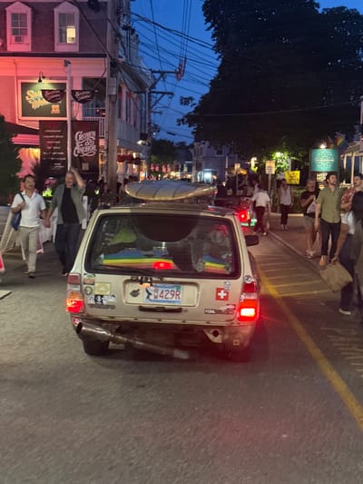 The image shows a street scene in Provincetown, Massachusetts, United States of America, during twilight hours. The focus is on a vintage, gold-colored Volvo station wagon with a surfboard strapped to its roof. The car is adorned with several stickers, including a Swiss flag and a license plate that reads "R 429R". Inside the car, colorful objects are visible through the rear window, suggesting personal belongings.

The car is parked on a street lined with buildings that appear to be shops and businesses, as indicated by signs like "Sailors Welcome!" and "Crown & Anchor". The buildings have illuminated windows and external lighting, casting a warm glow on the street.

Several people are present in the scene, walking on the sidewalk and in the street. They are casually dressed, suggesting a relaxed, possibly tourist-oriented atmosphere. The overall ambiance is that of a lively evening in a coastal town. The sky is a deep blue, transitioning from dusk to night.