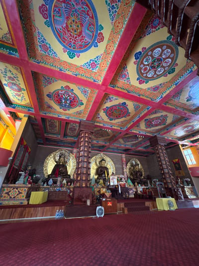 The image shows the interior of a Buddhist temple in Việt Trì, Vietnam. The ceiling is elaborately decorated with colorful paintings, featuring Buddhist motifs such as lotuses, mandalas, and mythical creatures like phoenixes. The walls are lined with small statues and religious artifacts. In the center of the room, three large golden statues of Buddha are prominently displayed on a raised platform. The floor is covered with a deep red carpet. The scene is well-lit, suggesting a daytime view. There are no people visible in the image. The overall impression is one of ornate religious devotion and peaceful contemplation.