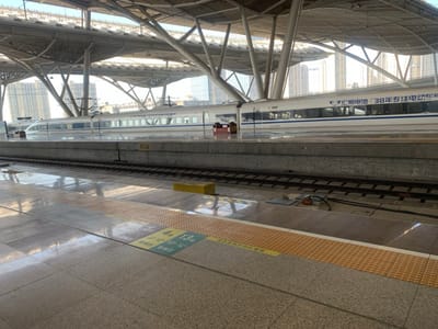 The image shows an empty train platform at Wuhan Railway Station in Wuhan, China, located at 30.608° N, 114.418° E. A white high-speed train is parked on the tracks to the right. The platform is covered by a large metal canopy supported by a complex network of steel beams. There are no people visible. The platform is made of grey tiles with a yellow stripe indicating the edge of the platform. The surface of the platform is wet, suggesting it may have recently rained. The train is labelled with the words "38 years of reform and opening up", which is a major political event in China. The platform is numbered 13 and the train is numbered 13 and 13. There is a yellow caution sign on the platform. The platform is bordered by a concrete wall, and the train is stopped several feet behind the wall. The station's architecture is modern, suggesting a relatively recent construction. A tall building with a glass facade can be seen in the background. The image is taken from the platform looking towards the train. The lighting suggests the image was taken during the day, but the time of day is unknown. The image is likely taken by a passenger waiting for a train.