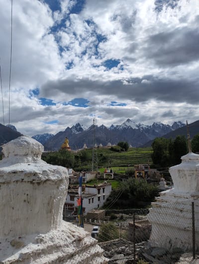 This is a photograph of the Karsha Monastery in Karsha, India, with a view of the surrounding landscape. In the foreground, to the left and right, are two whitewashed stupas, traditional Buddhist reliquaries. The stupa on the left is partially visible, showing its textured, weathered surface. The stupa on the right is more fully in view, with a chain-link fence in front of it.

In the middle ground, a village with white buildings and flat roofs is nestled in a green valley. Prayer flags in vibrant colors of red, blue, yellow, and green are strung between some of the buildings. Behind the village, a large, golden Buddha statue sits on a pedestal, overlooking the scene. Beyond the village and the Buddha statue, terraced fields climb the hillsides, dotted with trees.

In the background, a majestic mountain range with snow-capped peaks rises against a dramatic sky filled with white and grey clouds. A telecommunications tower is visible on a ridge to the right of the Buddha statue. A white car is parked on a road near the village. The overall impression is one of a remote, spiritual location set against a stunning natural backdrop.